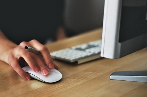 hand holding computer mouse with pen between fingers on a desk with a keyboard and monitor